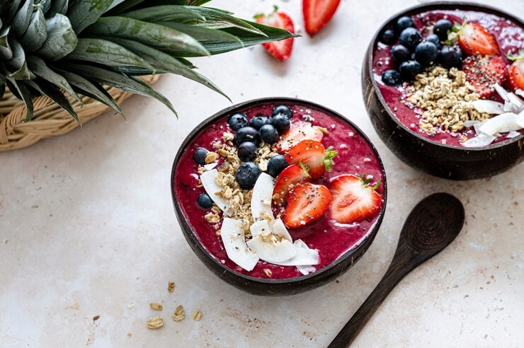 Two vibrant smoothie bowls, reminiscent of Vegan Delights, are topped with fresh strawberries, blueberries, granola, chia seeds, and coconut slices on a light surface. A wooden spoon and pineapple leaves accompany this acai bowl teeming with health benefits.