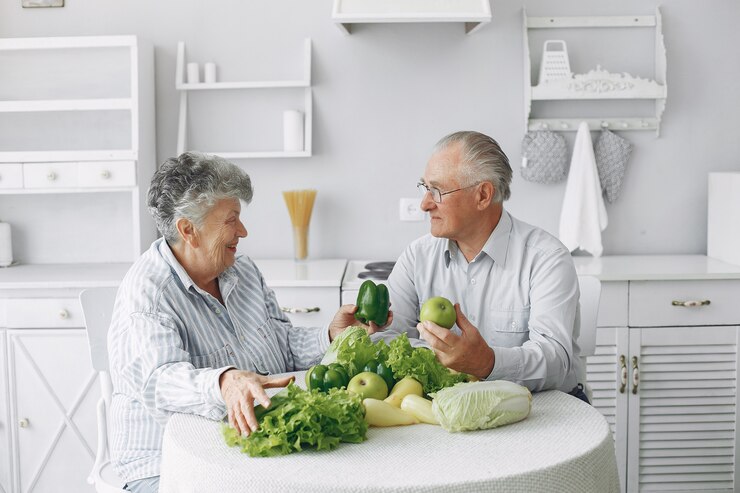 An elderly couple sits at a kitchen table covered with fresh vegetables, including lettuce and peppers, enjoying their vibrant vegan diet. They are smiling and engaged in conversation, embracing a lifestyle that supports Alzheimer's prevention. The kitchen has a light, minimalist design.