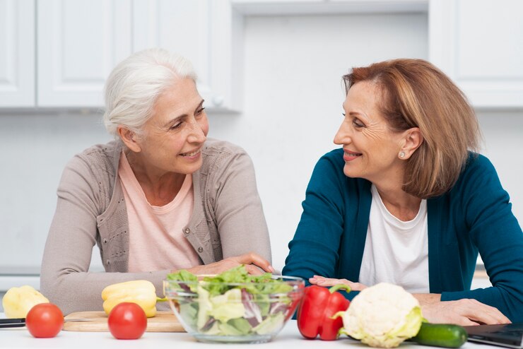 Two elderly women are smiling and chatting at a bright, white kitchen table with fresh vegetables like tomatoes, cucumbers, and peppers spread before them. A bowl of salad sits between the friends, hinting at their commitment to a vegan diet for Alzheimer's prevention.