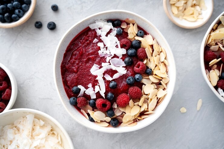 A vibrant acai bowl topped with fresh blueberries, raspberries, sliced almonds, and coconut flakes on a smoothie base, surrounded by small dishes of more berries and coconut on a light gray surface. Enjoy this vegan delight packed with health benefits in every bite.