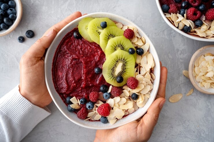 A person holds a vibrant acai bowl topped with sliced kiwi, blueberries, raspberries, and almond flakes. The background showcases more berries and almonds in small bowls, highlighting the vegan delights and health benefits packed in every colorful bite.