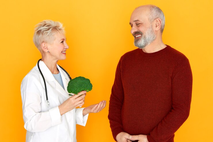 A doctor with short blonde hair, wearing a white coat and stethoscope, holds broccoli while smiling at a bearded man in a red sweater against a bright yellow background. She advocates for a vegan diet to prevent Alzheimer's.