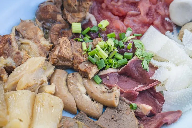 "An assortment of various cuts of raw and cooked meats, including tripe, liver, and sliced beef, garnished with chopped green onions and cilantro on a blue plate. The dish showcases a variety of textures and colours, highlighting different types of organ meats and muscle cuts."