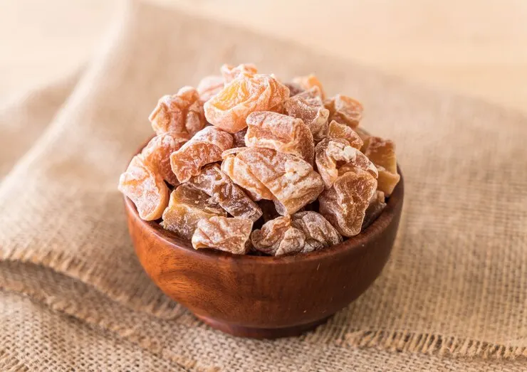 A bowl filled with peach gums, displayed on a wooden surface. The peach gums have a deep, rich colour and wrinkled texture, emphasising their preserved state and appealing appearance.