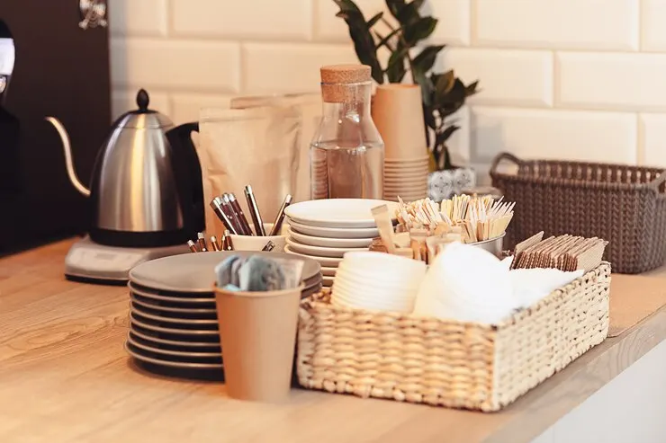 A cozy coffee station setup on a wooden countertop featuring stacked plates, cups, utensils, and a wicker basket holding disposable cups and stir sticks. A stainless steel electric kettle, a glass water bottle, and paper bags of coffee or tea are arranged neatly, with a small plant and woven storage basket adding a touch of decor in the background.