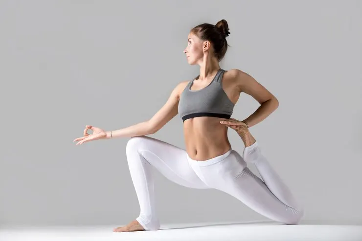 "Young woman in activewear performing a yoga pose on a grey background. She is in a low lunge position with one arm extended forward in a mudra and the other reaching back to hold her foot, displaying flexibility and balance." 