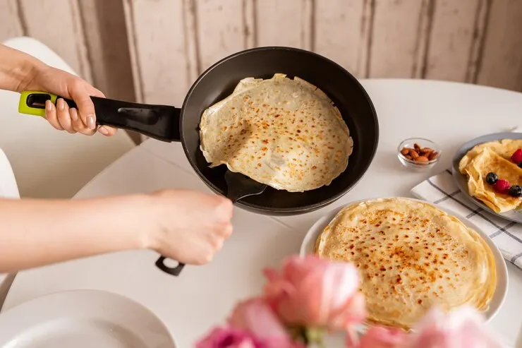 Hands young woman holding frying pan kitchen table while taking hot appetizing pancake put it top other crepes plate