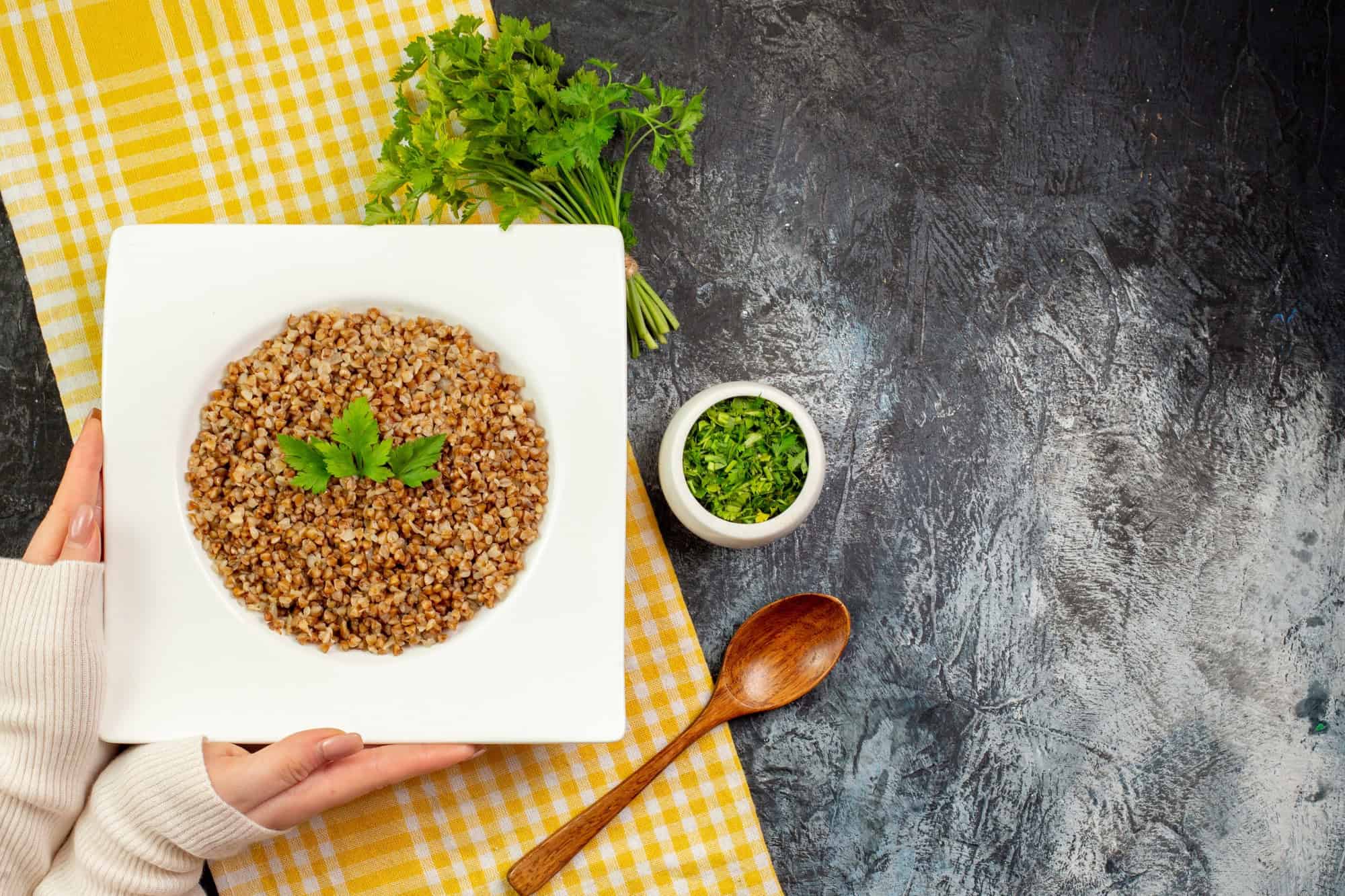 Top view of a bowl of cooked buckwheat garnished with parsley on a white plate, surrounded by fresh herbs and a wooden spoon on a rustic table
