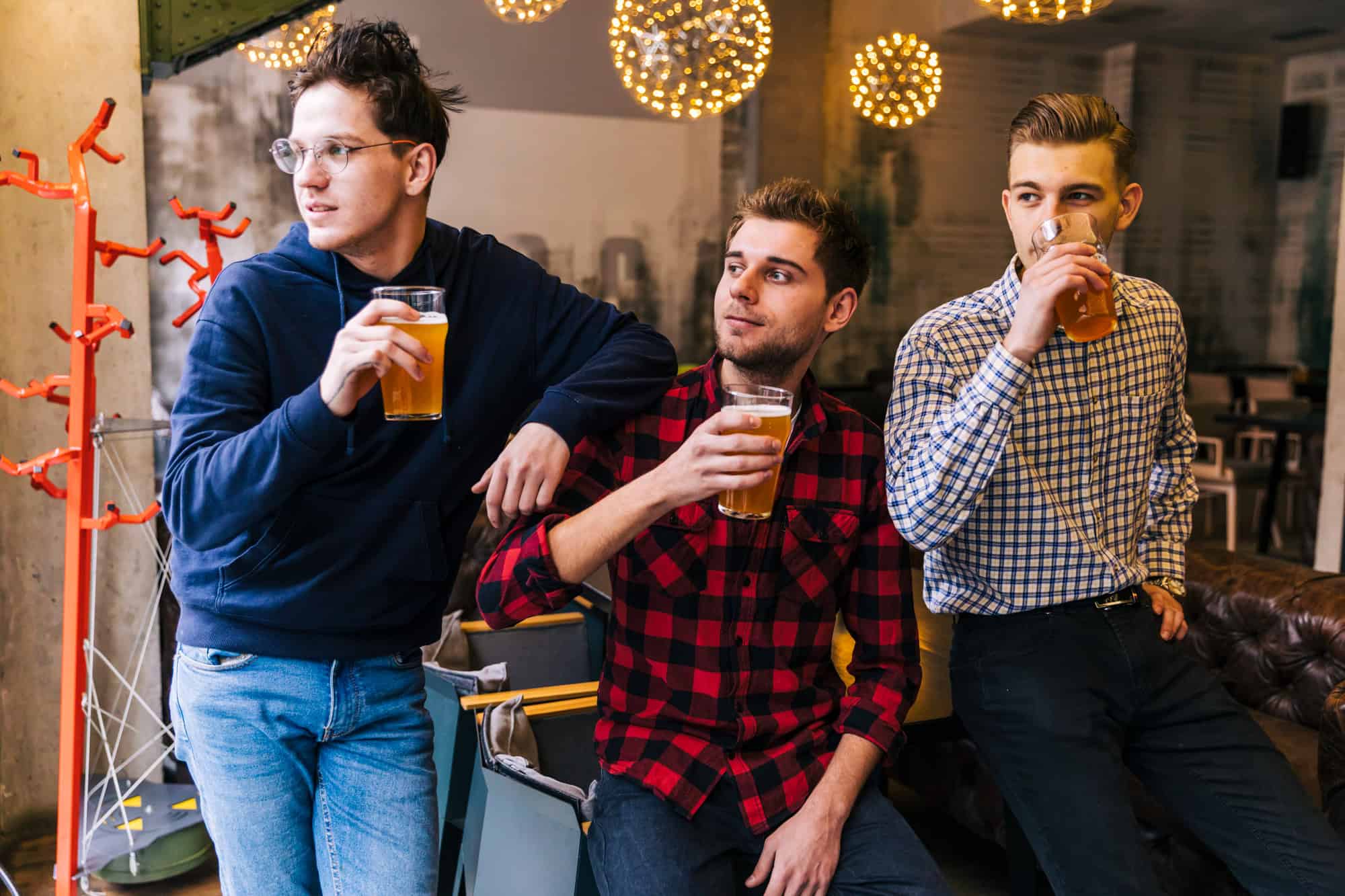 Three friends in casual clothing holding glasses of amber-coloured drinks in a cozy pub-style setting with warm hanging lights.