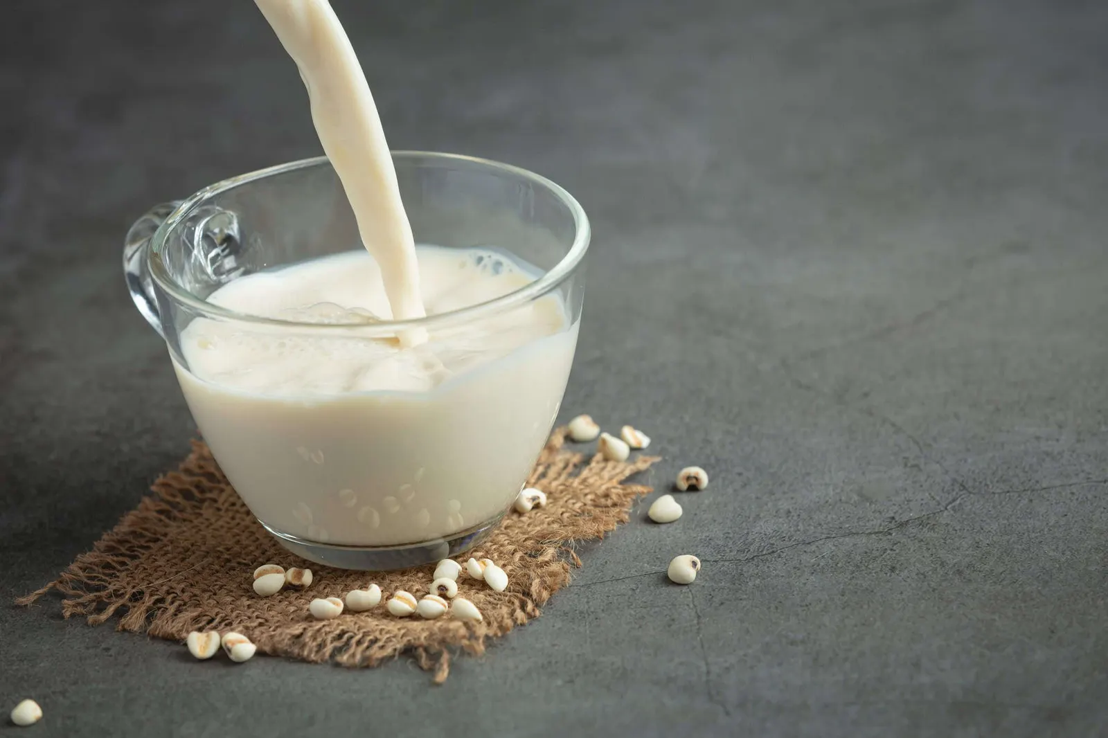 Soy milk or vegan whipping cream being poured into a clear glass cup, with scattered beans on a rustic cloth against a grey background.