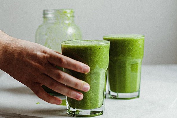 Two glasses of green smoothie on a white bench, one being picked up