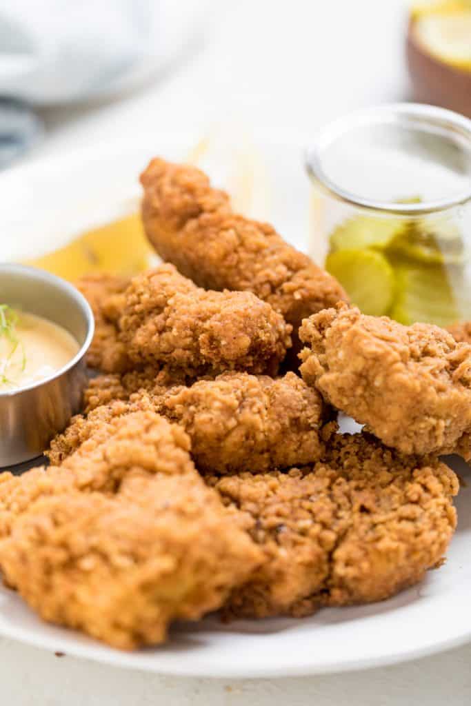 Vegan Chick-Fil-A Nuggets (CopyCat) A plate of golden-brown vegan Chick-Fil-A nuggets is served with lemon wedges, dill pickles in a jar, and a small dish of creamy sauce, all arranged on a white plate.