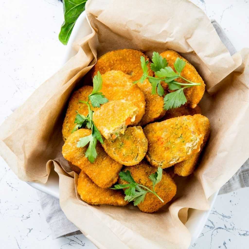 Chickpea Nuggets Recipe A white bowl filled with golden-brown, breaded chickpea nuggets, garnished with fresh parsley. The bowl is lined with brown parchment paper, emphasizing this delightful veggie nugget recipe as it rests on a white, textured surface.