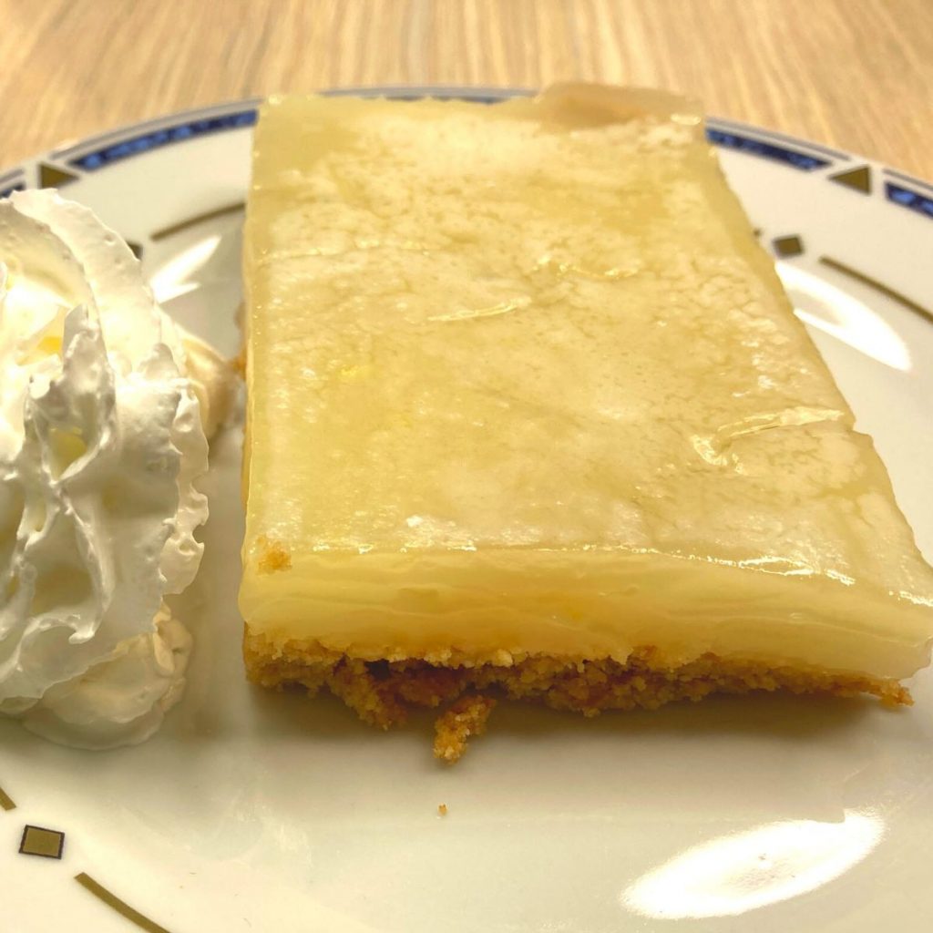 Creamy Vegan Lemon Bars A slice of creamy vegan lemon bars with a translucent top layer sits on a crumbly base, gracing a white plate with a decorative blue and gold rim. Beside it rests a dollop of whipped cream, all set against a light wooden background.