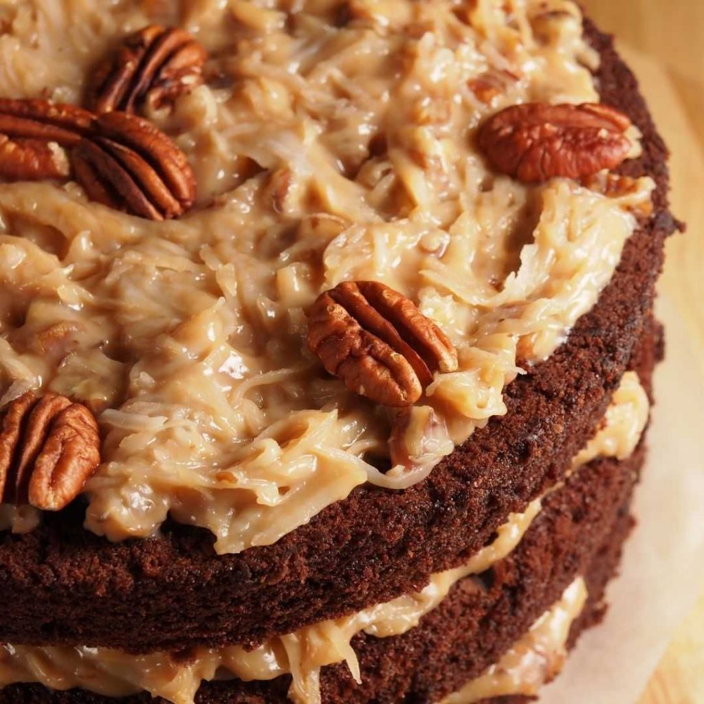 Close-up of a vegan German chocolate cake with layers of rich chocolate sponge and caramelized coconut pecan frosting. Whole pecans adorn the top, adding a decorative touch.
