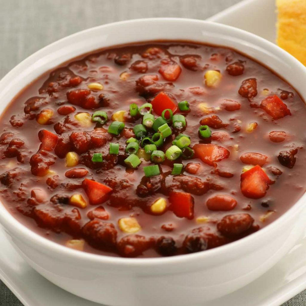 A bowl of vegan chili, inspired by a Walt Disney copycat recipe, is topped with chopped green onions. It features kidney beans, corn, and tomato chunks in a white bowl on a white plate. Cornbread peeks from the side, completing this delightful dish.