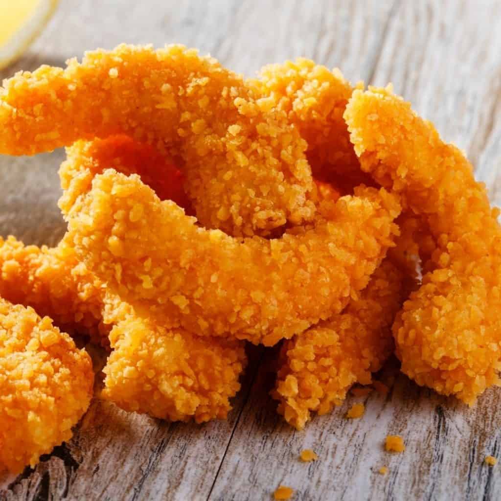 Close-up of crispy, breaded vegan shrimp piled on a rustic wooden surface. The golden-brown coating is flaky, suggesting a crunchy texture. A hint of lemon is visible in the background, adding a touch of color and freshness to the scene.