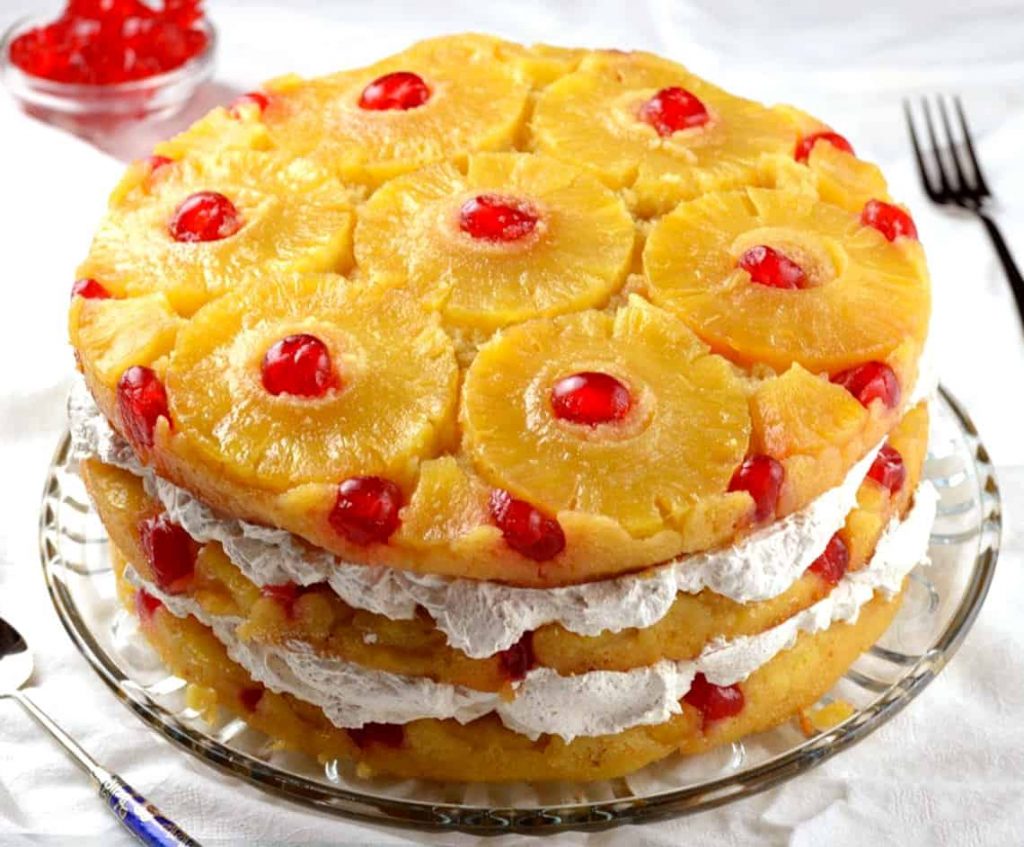 Vegan Pineapple Upside-Down Cake Recipe A delicious vegan pineapple upside-down cake with whipped cream and maraschino cherries, beautifully arranged on a clear glass plate.