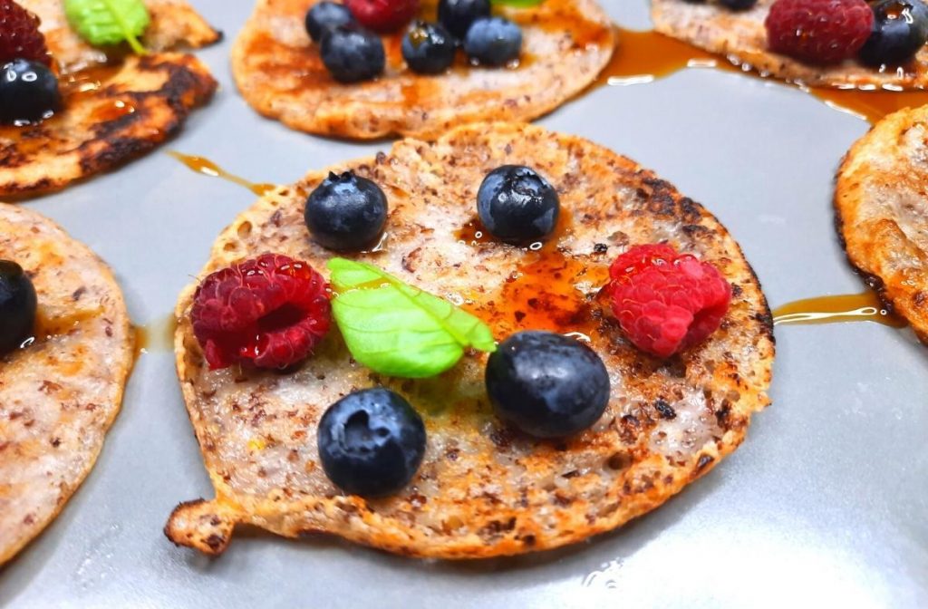 Close-up of vegan tapioca flour pancakes topped with blueberries, raspberries, and a drizzle of syrup, garnished with mint leaves. The pancakes sit on a gray plate, showcasing a delightful mix of colors and textures.