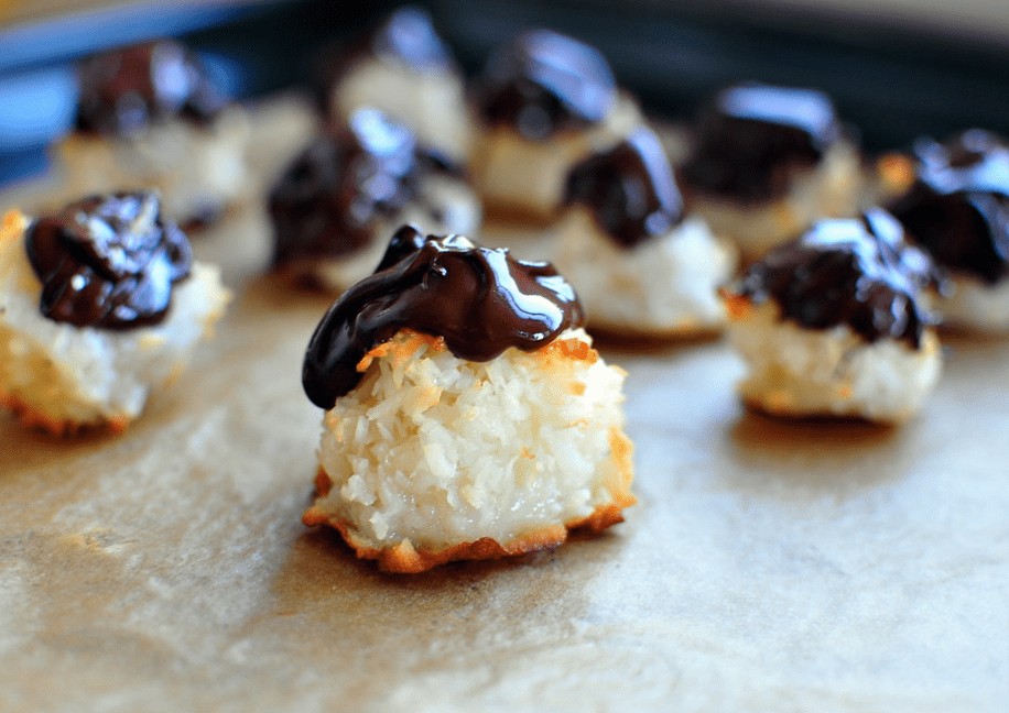Vegan Coconut Macaroons (Easy & Tasty Recipe) Close-up of vegan coconut macaroons topped with melted dark chocolate. They are placed on a parchment-lined baking sheet, with a blurred background showing more macaroons.