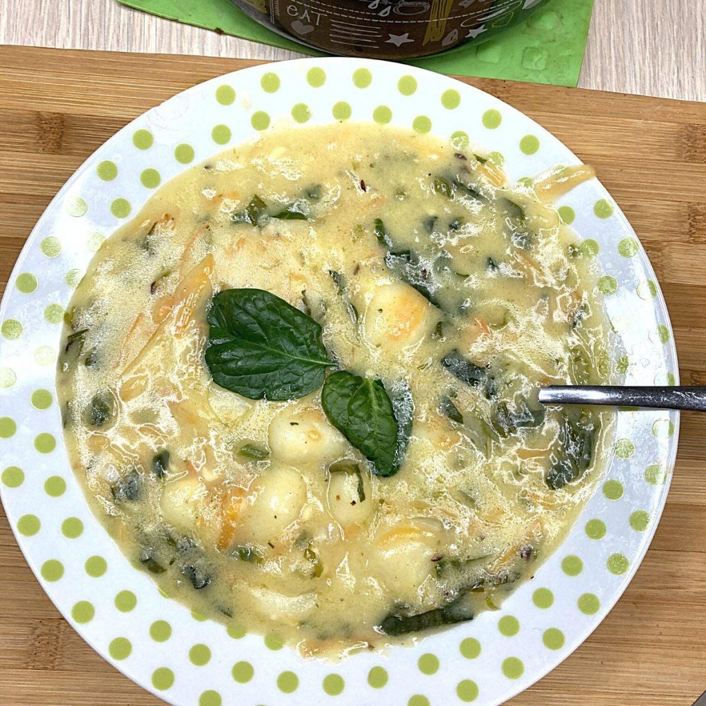 A bowl of creamy vegan gnocchi soup with spinach and shredded carrots, garnished with a sprig of fresh basil. The soup is served on a white plate with green polka dots, resting on a wooden surface.