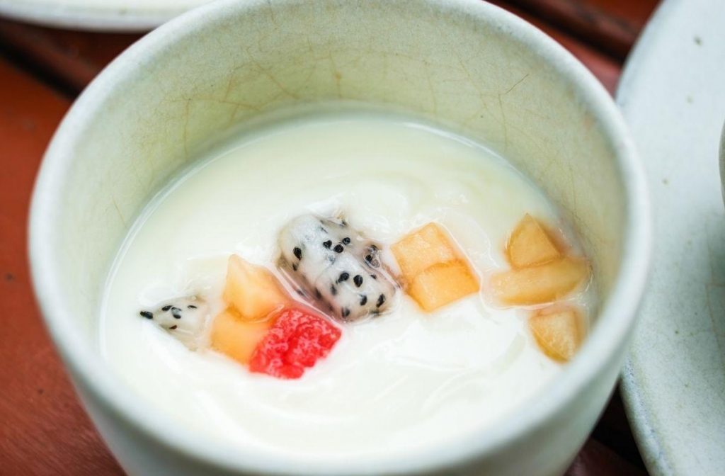 Easy Almond Tofu Recipe A bowl of creamy yogurt topped with small pieces of dragon fruit, melon, and a single red berry evokes the delightful contrast seen in almond tofu. The colorful fruits add vibrant contrast to the pale yogurt, creating an appetizing and visually appealing dish.