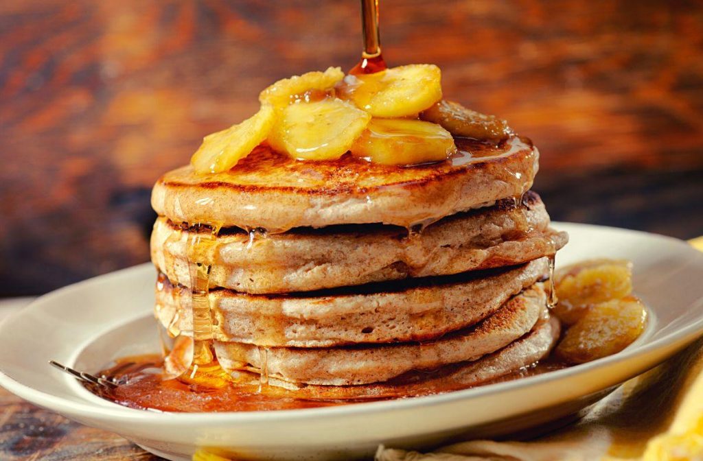 Easy AIP Banana Pancakes Recipe A stack of delicious AIP banana pancakes is topped with sliced bananas and drizzled with syrup on a white plate. The background's warm wooden tone creates a cozy breakfast atmosphere.