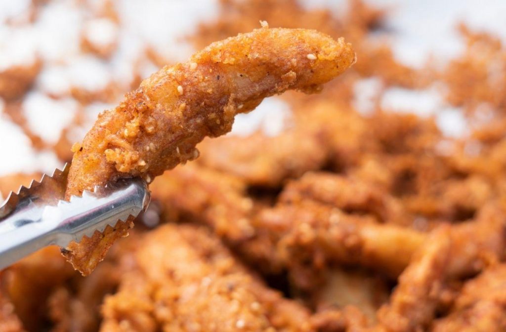 Banana Blossom Recipe (Vegan Fish & Chips) A close-up image of a golden-brown fried chicken piece, reminiscent of a banana blossom recipe, held by metal tongs. The crispy coating is textured and speckled, with more pieces of this delicious creation blurred in the background.