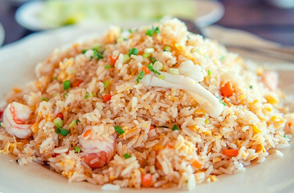A vegan hibachi-inspired plate of fried rice brimming with vegetables. The dish features an array of carrots and green onions blended seamlessly into the rice, while a background of blurred sliced cucumbers adds a touch of freshness.