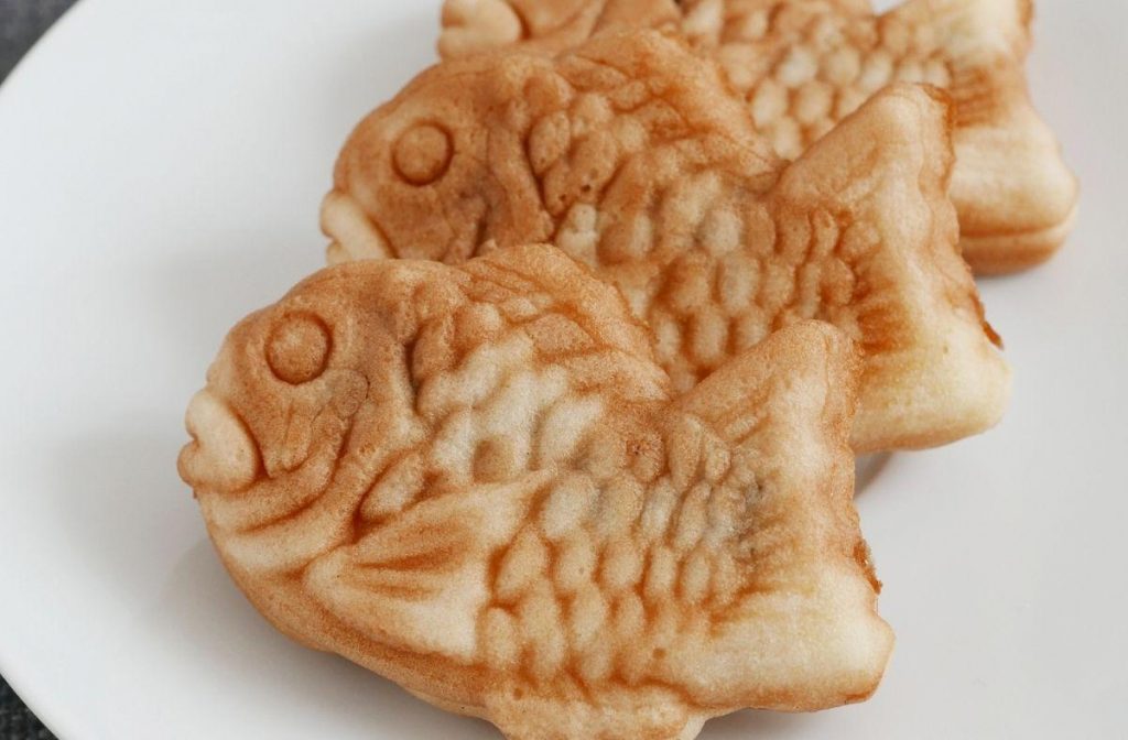Vegan Taiyaki Recipe (Japanese Fish Waffle) Three vegan taiyaki pastries are neatly arranged on a white plate. Each fish-shaped pastry features detailed scales and fins, boasting a golden-brown color that beautifully resembles traditional taiyaki.