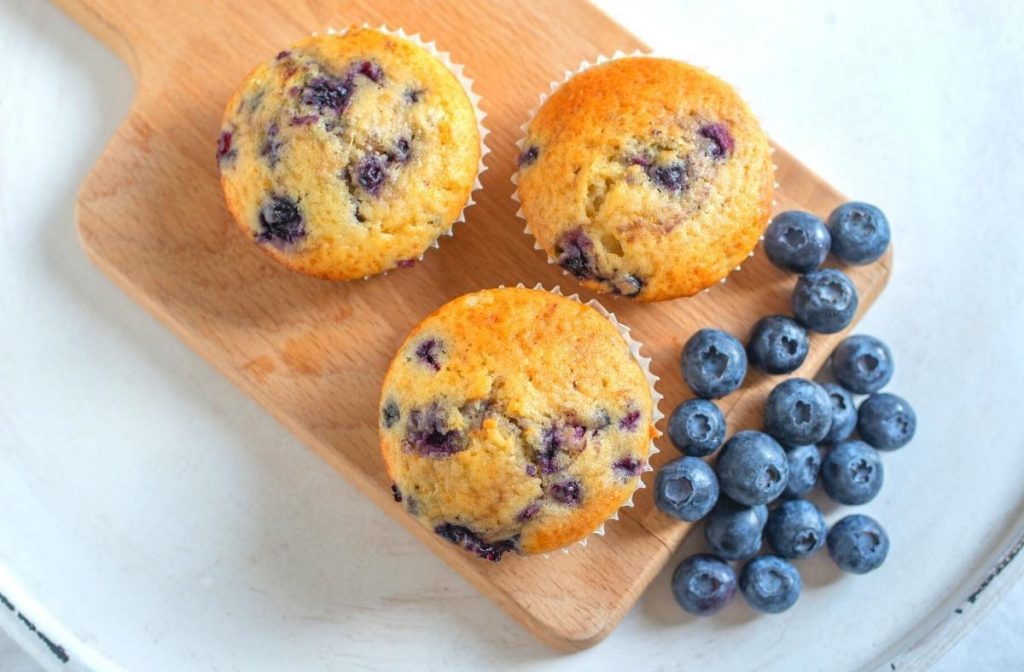 Three vegan blueberry muffins rest on a wooden board, accompanied by a handful of fresh blueberries. The muffins are golden brown with visible blueberries baked inside, set on a light-colored background.