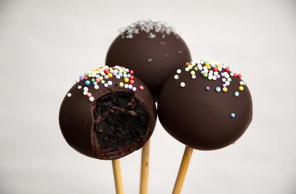 Easy and Tasty Vegan Cake Pops Recipe Three vegan chocolate cake pops on sticks, each coated in a smooth layer of dark chocolate. Two are decorated with colorful sprinkles, while the third has silver sprinkles. One of them has a bite taken out, revealing a rich, dark interior.