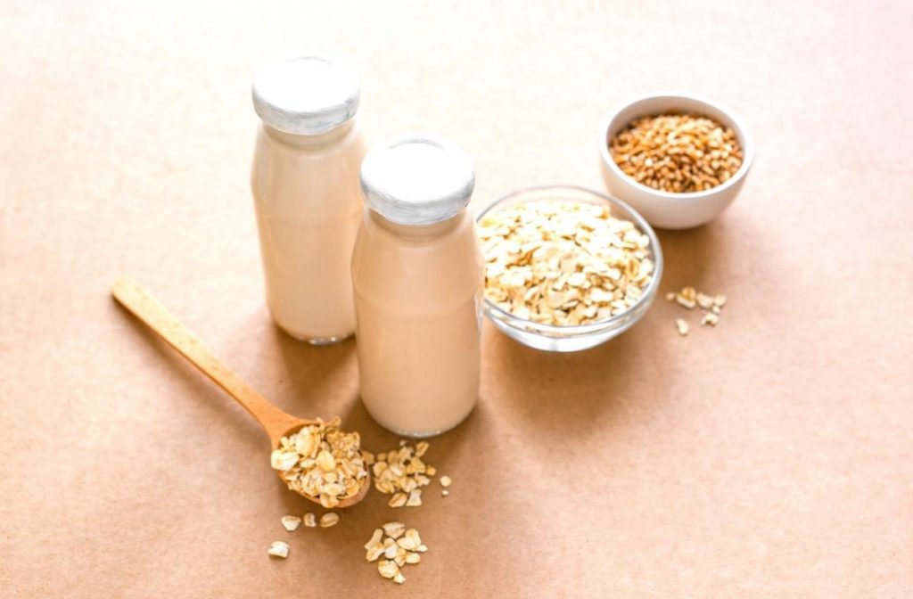 How to make Oat Milk! (Easy & Quick) Two glass bottles of oat milk are placed on a brown surface, inviting you to explore how to make oat milk. Nearby are two bowls: one brimming with whole oats and another with oat flakes. A wooden spoon filled with oat flakes completes the rustic arrangement.