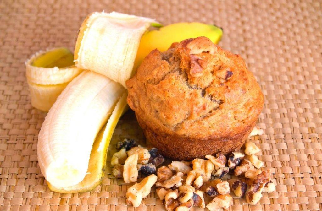 A vegan banana nut muffin sits on a woven mat alongside a peeled banana and scattered walnuts.