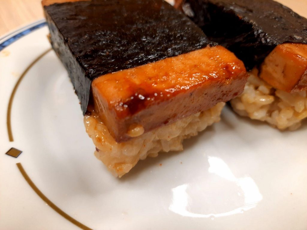 Vegan Tofu “SPAM” Musubi Recipe Close-up of two pieces of musubi on a plate, each featuring a glazed tofu slice on rice and wrapped with seaweed. The musubi recipe shines on a plate adorned with a simple geometric pattern near the edge.