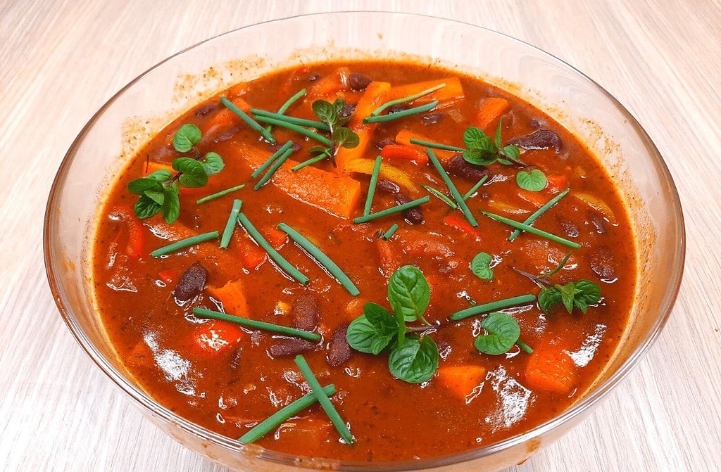 A bowl of hearty vegetable stew with dark beans, orange carrots, and chunks of potatoes in a rich, red chakalaka sauce. Garnished with green herbs and chives, it sits on a light wooden table.