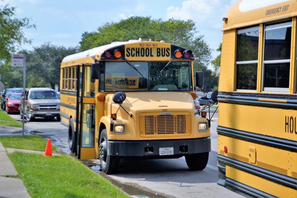 Impossible Foods Puts New Program on Sustainability for the K-12 Curriculum Two yellow school buses, staples of the K-12 curriculum commute, are parked on a suburban street. One is partially visible on the right, while the other faces forward. Cars line the curb, with a small orange cone near one bus. Lush trees provide a backdrop hinting at sustainability in design.
