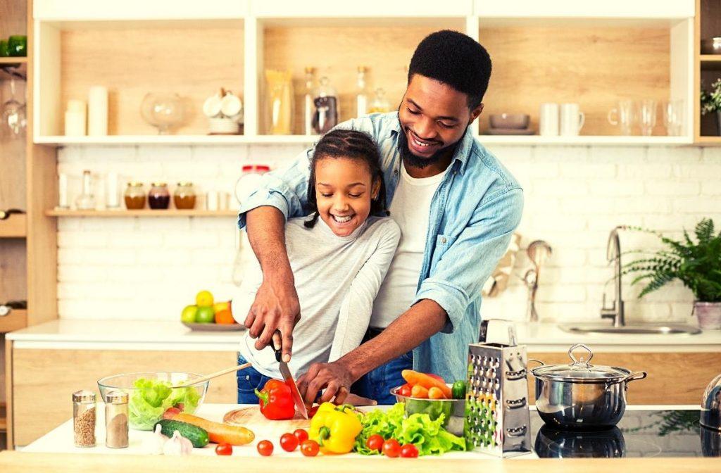 In a modern kitchen with white and wooden accents, a man and a child smile as they prepare a meal for Meat-Free Friday. They're chopping vegetables like peppers, tomatoes, and lettuce amidst the wonderful aroma of fresh ingredients, embracing both culinary traditions and sustainability.