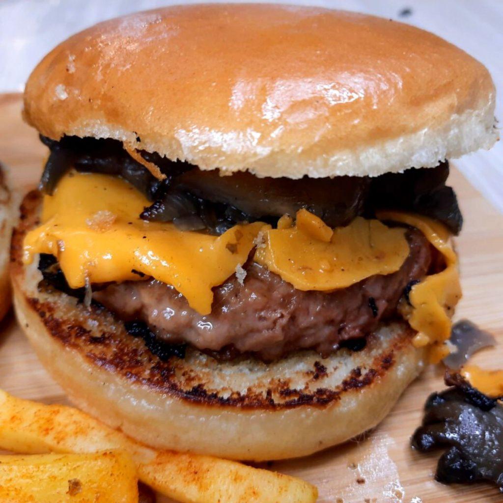 Close-up of a Vegan Mushroom Swiss Burger featuring a juicy plant-based patty, melted Swiss cheese, grilled mushrooms, and sauce, all sandwiched in a shiny, toasted bun. Accompanied by a side of seasoned fries on a wooden surface.