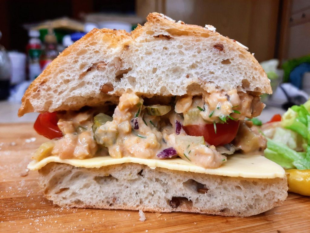 A close-up of a sandwich with a thick slice of bread, filled with cheese, cherry tomatoes, a creamy vegan tuna filling with herbs, and a side of leafy greens. The background is a blurred kitchen scene.