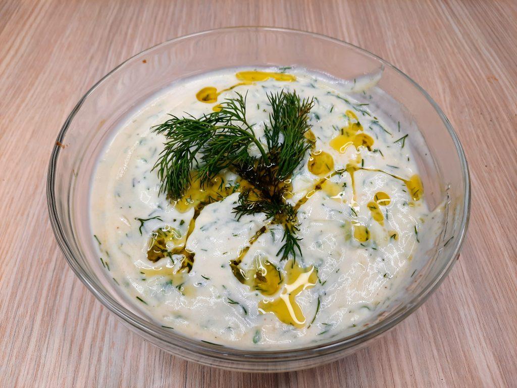 A glass bowl brimming with creamy vegan tzatziki sauce, garnished with a sprig of fresh dill and drizzled with olive oil, rests elegantly on a wooden surface.