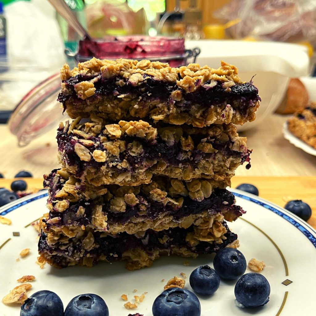 Blueberry Jam Oatmeal Bars Recipe (Gluten-Free) A stack of three delicious blueberry jam oatmeal bars sits on a white plate surrounded by fresh blueberries. These bars feature a crumble topping with visible layers of oats and fruit. In the background, there's a jar of blueberry jam and a bowl, enhancing the delightful scene.