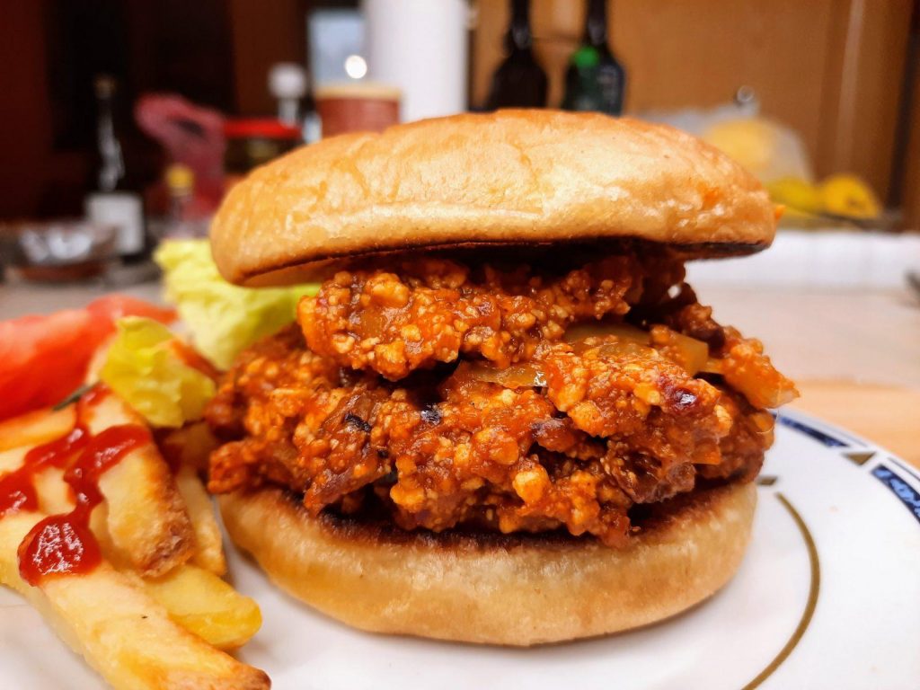 A close-up of a delicious vegan sloppy joe with a thick, crumbly filling served on a bun. Accompanied by lettuce, a tomato slice, and ketchup-covered fries on a white plate, the background reveals condiments on a wooden surface.