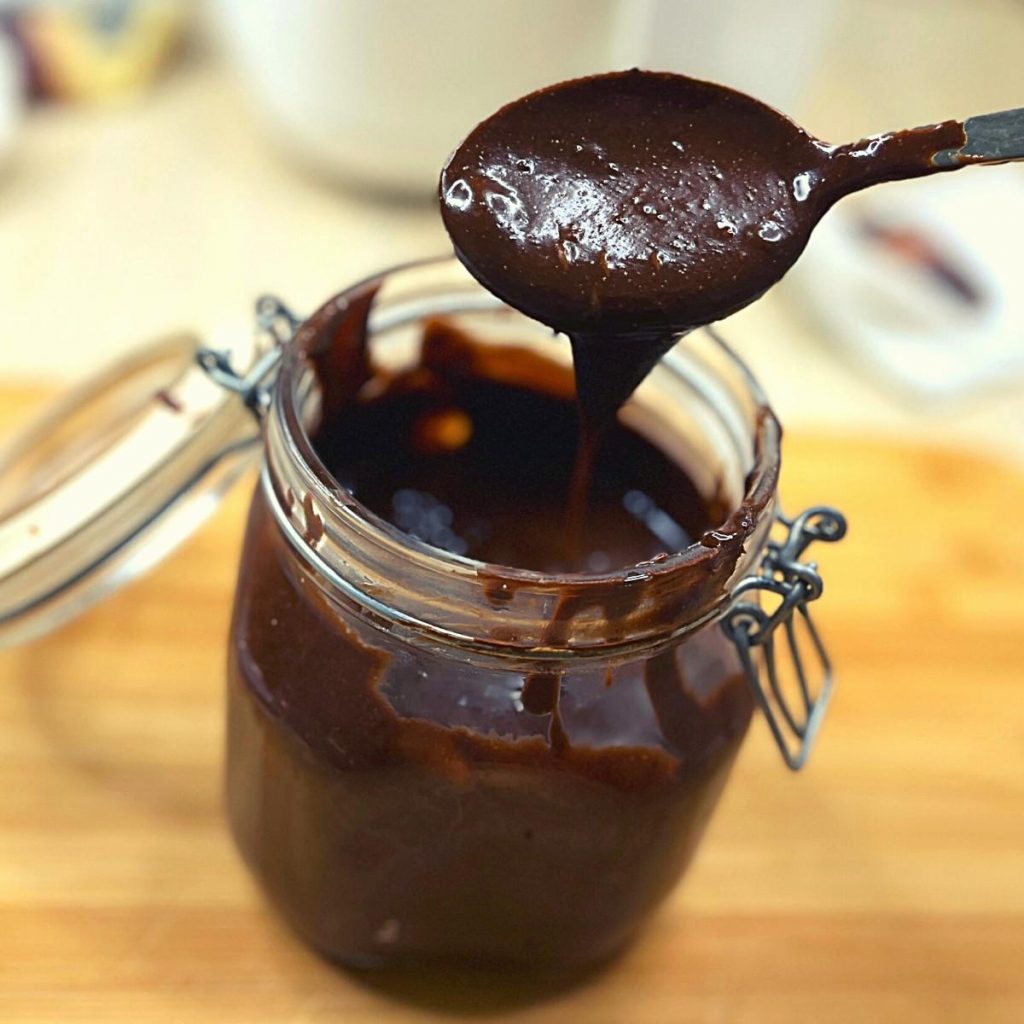 Homemade Vegan Nutella Recipe & Store Bought Alternatives A glass jar filled with rich, dark vegan Nutella sauce sits on a wooden surface. A spoon lifts the thick sauce from the jar, catching light reflections on its smooth texture. The jar, slightly open, features a metal clasp.