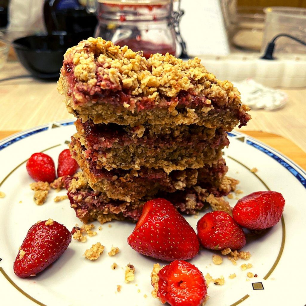 Irresistible Strawberry Jam Oatmeal Bars (Gluten-Free) A stack of crumbly strawberry jam oatmeal bars sits on a white plate, surrounded by fresh strawberries. The background reveals a blurred kitchen scene with various utensils and ingredients, capturing the essence of homemade comfort.