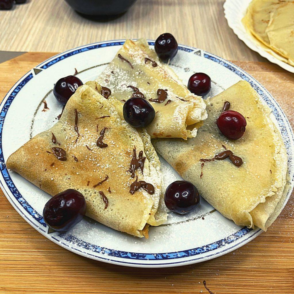 A plate of folded vegan crepes garnished with cherries and drizzled with chocolate sits on a wooden surface. A dusting of powdered sugar adds a sweet touch to the crepes and plate. Another plate with crepes is partially visible in the background, showcasing this delightful plant-based treat.