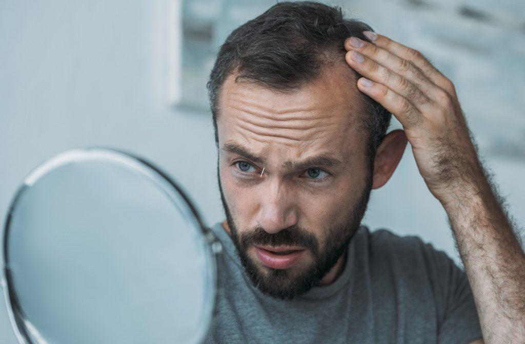 A Comprehensive Guide to Stress-Induced Hair Loss and Its Treatments A man with a beard looks at his reflection in a handheld mirror, touching his hair with concern. Stress-induced hair loss seems to weigh on him. He wears a gray shirt, and the background is softly blurred, emphasizing his focused worry.