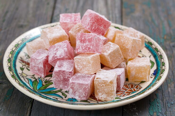 A decorative plate filled with colorful cubes of powdered Turkish delight in pink and yellow, crafted from an authentic Turkish Delight recipe, rests elegantly on a rustic wooden surface.