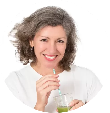 Trish-Tucker-May-1-png-420x429 A person with shoulder-length gray hair, a true veganiac, smiles while sipping a green beverage through a straw. They are wearing a white shirt, perfectly complementing the pure, minimalist white background.