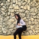 A person in a striped shirt and black pants stands smiling against a stone wall, radiating veganiac vibes as they hold a black bag and a small item in their hand.